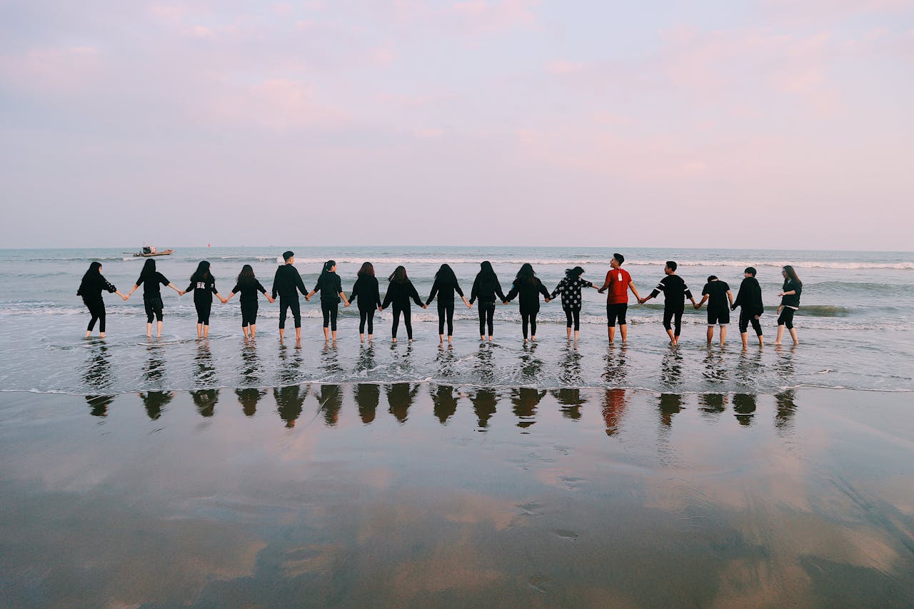 our-story-01 A large group holding hands at the beach during sunset, reflecting in the water.